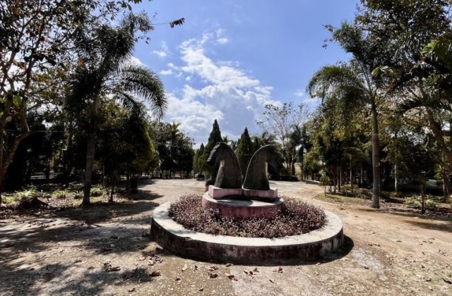 Two dark horse-head sculptures stand in a circular planter with reddish plants, surrounded by green trees and dirt paths under a blue, cloudy sky.