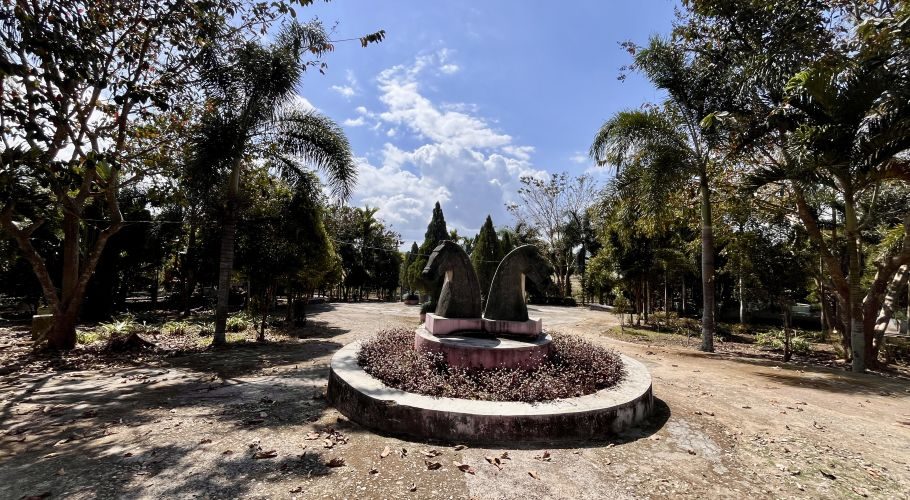 Two dark horse-head sculptures stand in a circular planter with reddish plants, surrounded by green trees and dirt paths under a blue, cloudy sky.