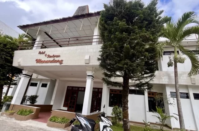 The white entrance of Hotel Manandang is framed by tall green trees, with two white motorbikes parked in front under a cloudy sky.