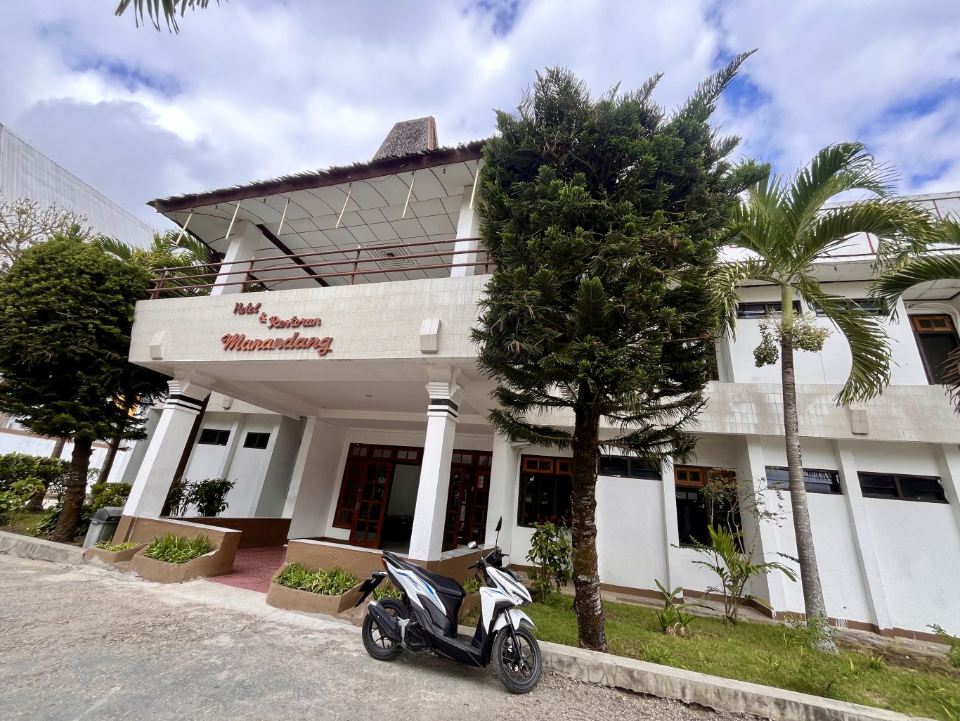 The white entrance of Hotel Manandang is framed by tall green trees, with two white motorbikes parked in front under a cloudy sky.