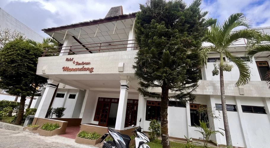 The white entrance of Hotel Manandang is framed by tall green trees, with two white motorbikes parked in front under a cloudy sky.