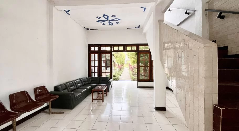 Manandang Hotel lobby features a black sofa, brown chairs, white tiled floor, and a patterned ceiling, opening to a lush garden pathway.