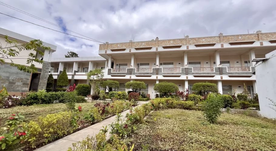 Manandang Hotel's light-colored building with balconies overlooks a tranquil garden with a winding pathway and diverse plants.