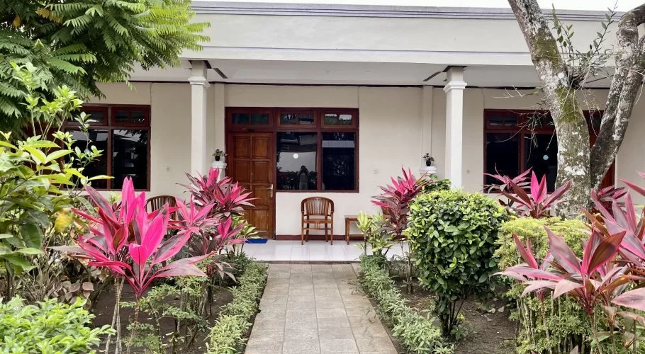 A cream-colored hotel building with a dark wooden door and windows, fronted by a paved path and vibrant pink and green tropical plants.
