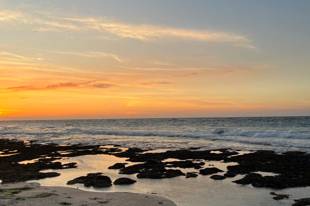 Sunset over a beach with an orange and yellow sky. Waves crash on the dark rocky foreground, reflecting the sky's warm colors.