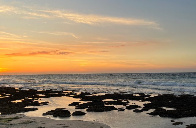 Sunset over a beach with an orange and yellow sky. Waves crash on the dark rocky foreground, reflecting the sky's warm colors.