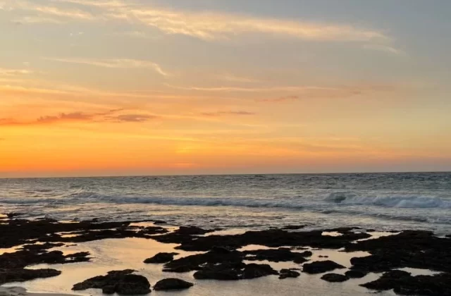 Sunset over a beach with an orange and yellow sky. Waves crash on the dark rocky foreground, reflecting the sky's warm colors.