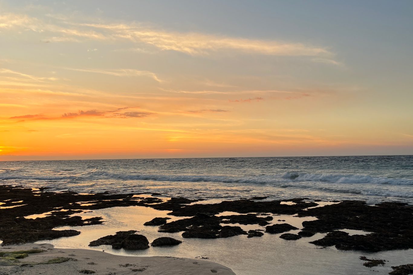 Sunset over a beach with an orange and yellow sky. Waves crash on the dark rocky foreground, reflecting the sky's warm colors.
