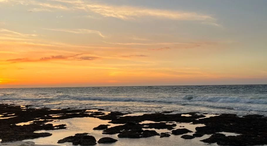 Sunset over a beach with an orange and yellow sky. Waves crash on the dark rocky foreground, reflecting the sky's warm colors.