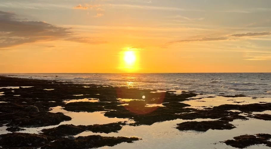 The bright orange sun sets over the ocean, casting a warm glow on the sky and reflecting in tidal pools among dark rocks on Mananga Aba Beach.