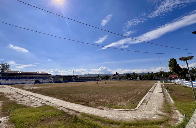 Manda Elu Stadium in Waikabubak features a large, dry grass football field, blue and white grandstands, and a bright blue sky with white clouds.