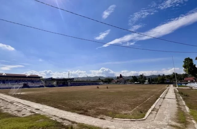 Manda Elu Stadium in Waikabubak features a large, dry grass football field, blue and white grandstands, and a bright blue sky with white clouds.