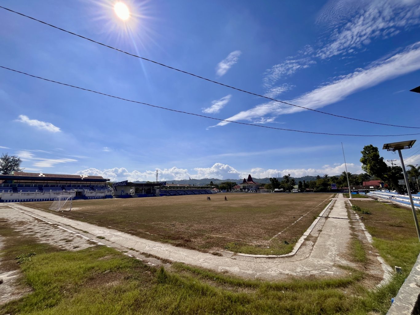 Manda Elu Stadium in Waikabubak features a large, dry grass football field, blue and white grandstands, and a bright blue sky with white clouds.
