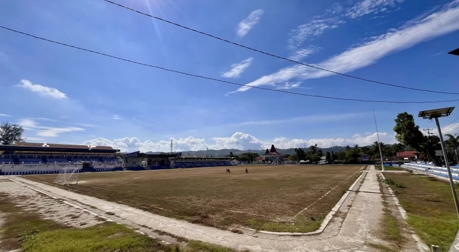 Manda Elu Stadium in Waikabubak features a large, dry grass football field, blue and white grandstands, and a bright blue sky with white clouds.