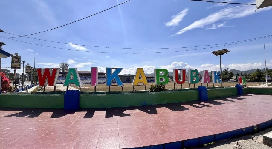 A large, colorful sign spelling WAIKABUBAK stands on a green base in front of Manda Elu Stadium under a bright blue sky.