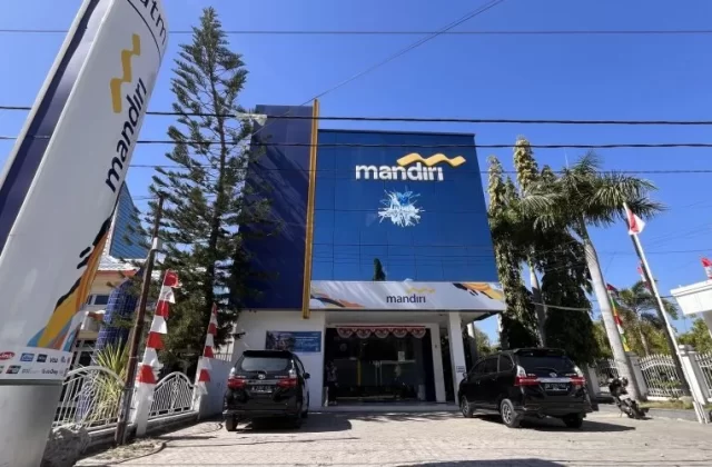 A modern blue and white Mandiri Bank branch building stands prominently under a clear blue sky, with two black cars parked in front and a tall bank sign on the left.