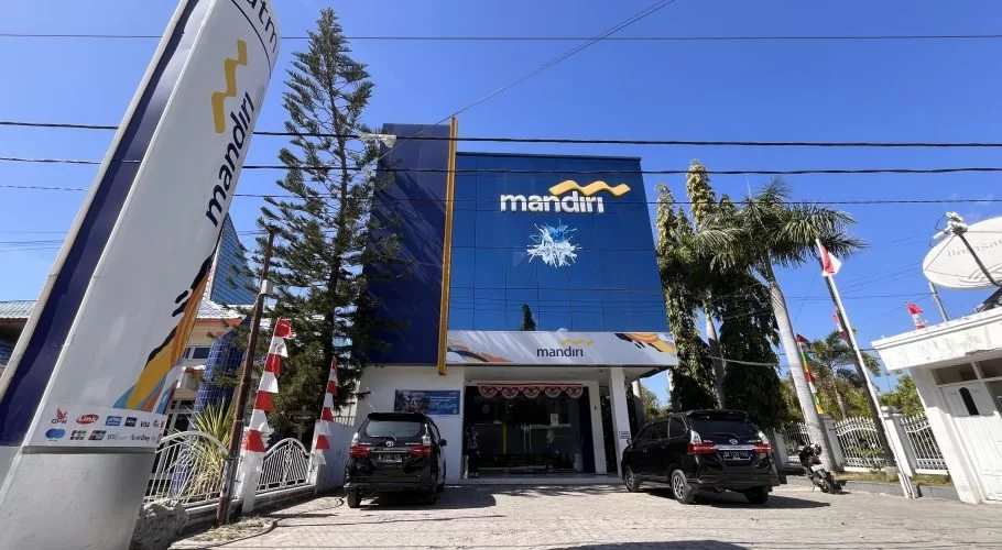 A modern blue and white Mandiri Bank branch building stands prominently under a clear blue sky, with two black cars parked in front and a tall bank sign on the left.