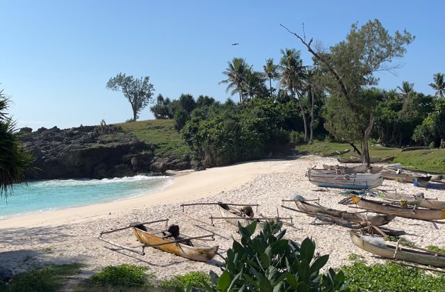 A pristine white sand beach with turquoise water is framed by rocky cliffs and lush green vegetation under a clear blue sky, with several traditional wooden outrigger boats pulled ashore.