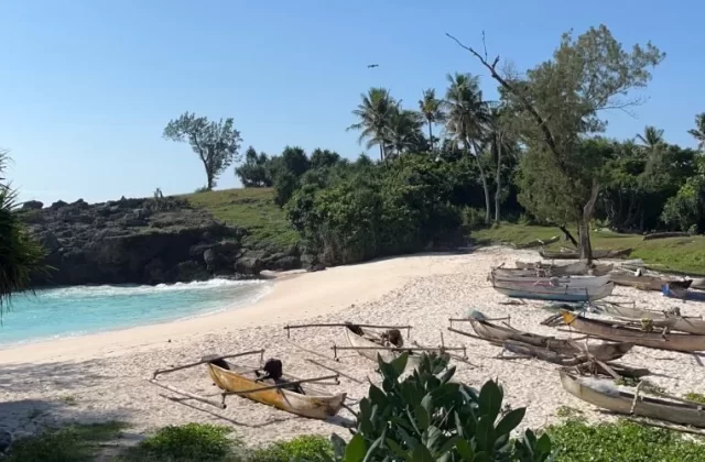A pristine white sand beach with turquoise water is framed by rocky cliffs and lush green vegetation under a clear blue sky, with several traditional wooden outrigger boats pulled ashore.