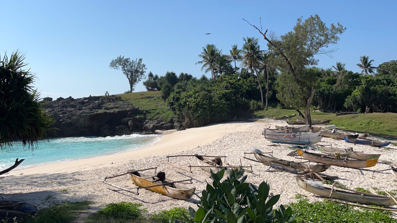 A pristine white sand beach with turquoise water is framed by rocky cliffs and lush green vegetation under a clear blue sky, with several traditional wooden outrigger boats pulled ashore.