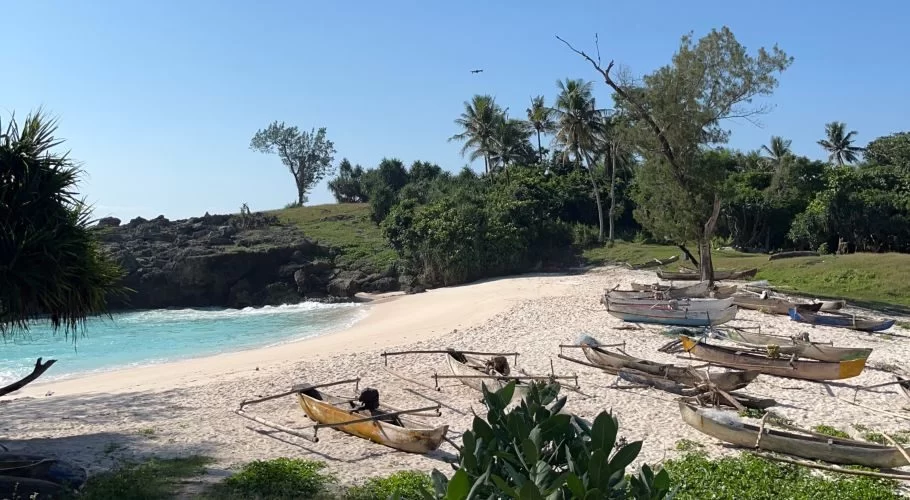 A pristine white sand beach with turquoise water is framed by rocky cliffs and lush green vegetation under a clear blue sky, with several traditional wooden outrigger boats pulled ashore.