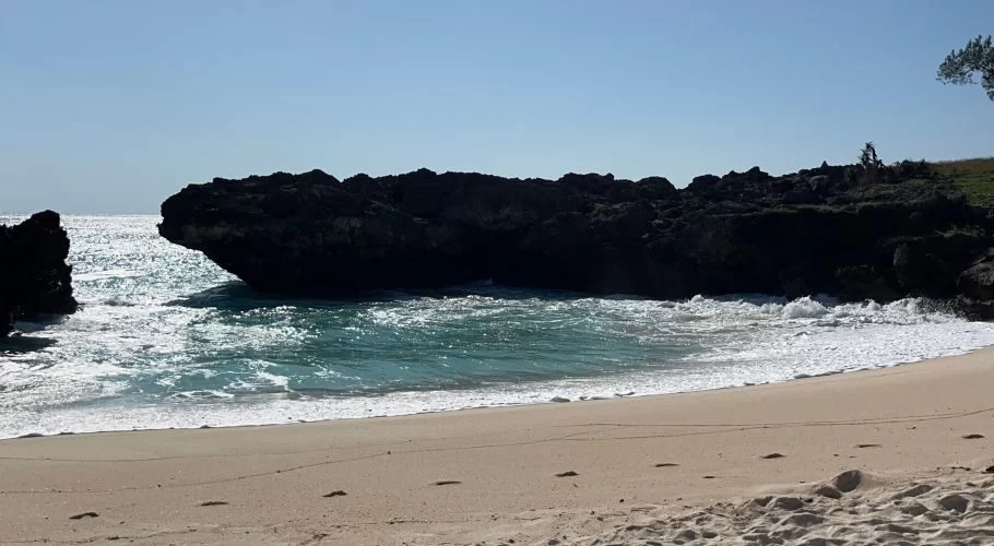 Mandorak Beach features light sand, clear turquoise waves breaking on the shore, and dark, rugged rock formations under a bright blue sky.
