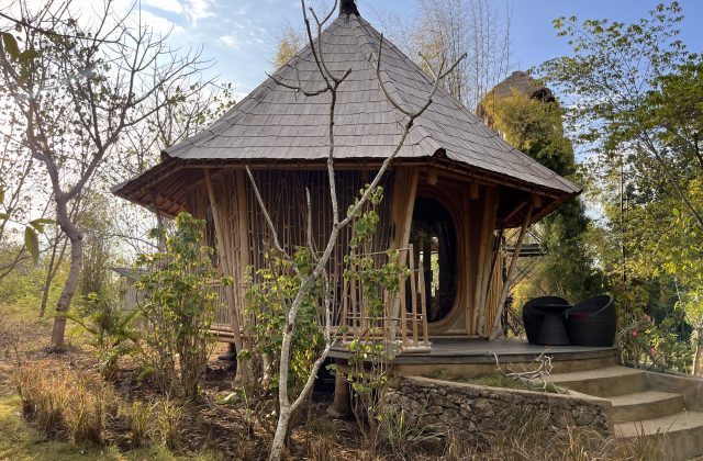 A round, light brown bamboo pavilion, part of Maringi Hotel, stands amidst dry trees and green foliage under a bright sky.
