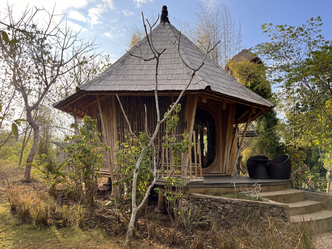A round, light brown bamboo pavilion, part of Maringi Hotel, stands amidst dry trees and green foliage under a bright sky.
