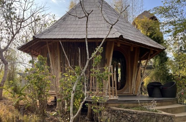 A round, light brown bamboo pavilion, part of Maringi Hotel, stands amidst dry trees and green foliage under a bright sky.