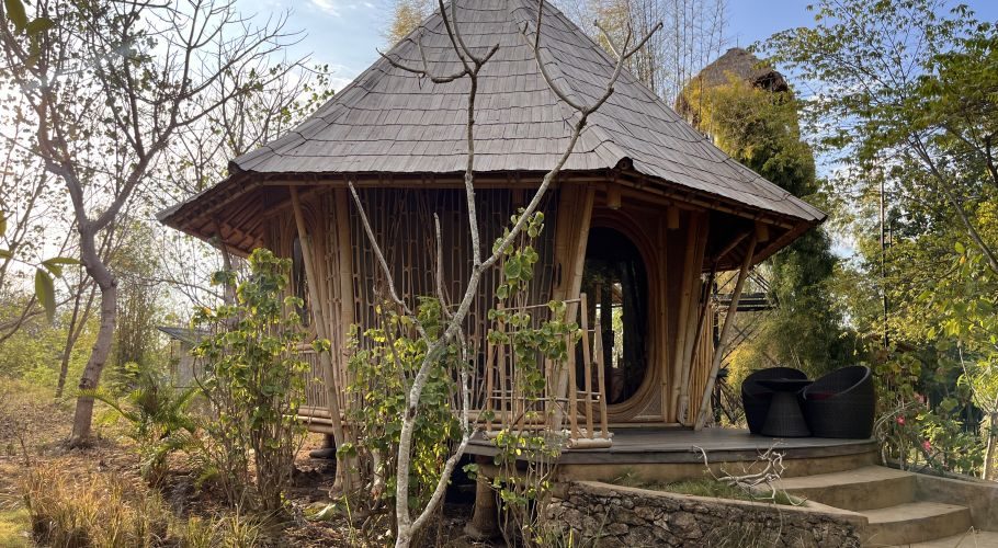 A round, light brown bamboo pavilion, part of Maringi Hotel, stands amidst dry trees and green foliage under a bright sky.