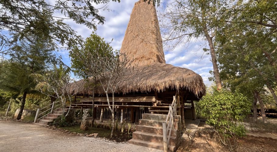 A traditional Sumba-style building with a tall, conical thatched roof and bamboo structure stands amidst green trees under a partly cloudy sky.