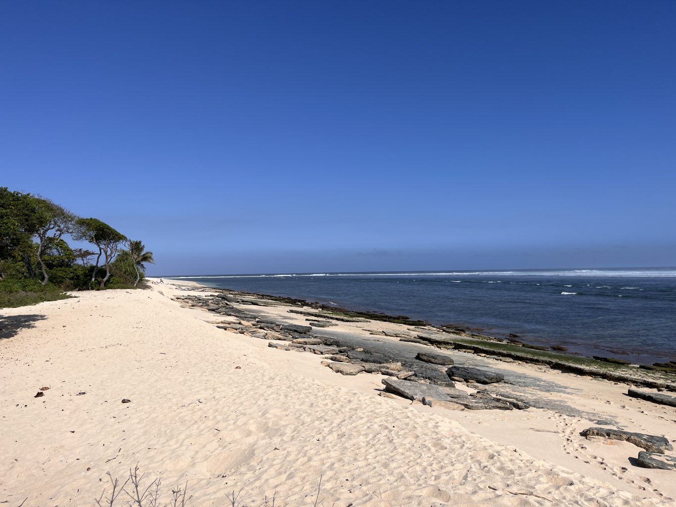 A wide white sand beach stretches along a deep blue ocean with distant breaking waves under a clear blue sky, bordered by green trees and dark coral formations.
