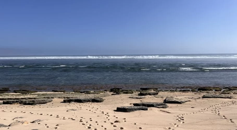Light brown sand with footprints and dark rocks leads to clear dark blue ocean waves under a bright blue sky at Marosi Beach.