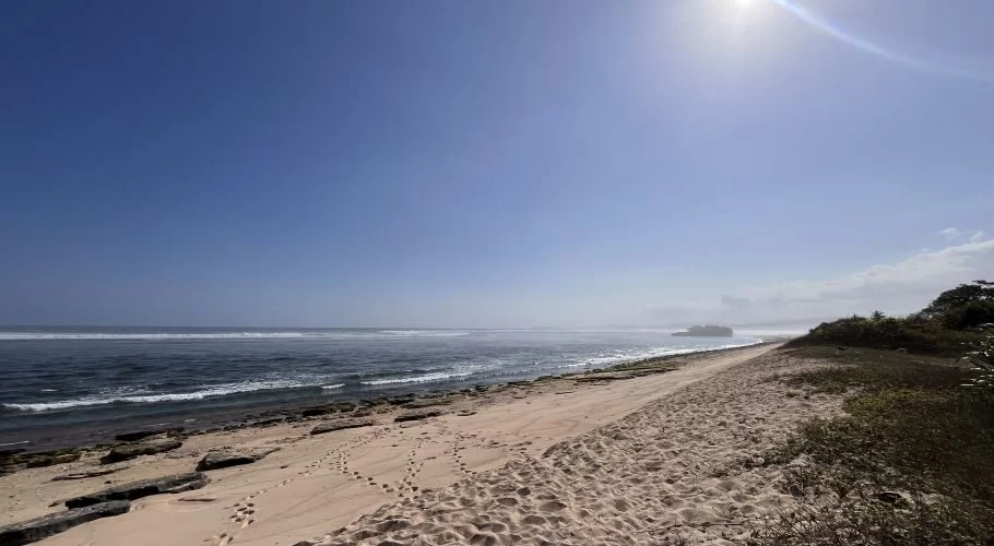 A wide, pristine white sand beach stretches along a blue ocean with gentle waves under a bright blue sky, showing footprints and some dark rocks near the waterline.
