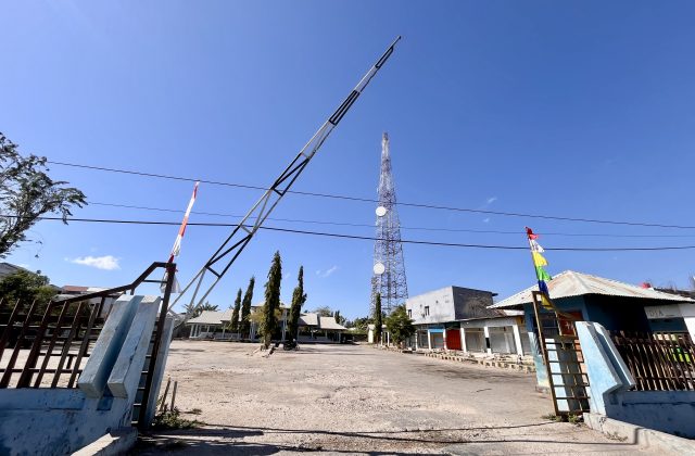 The entrance to Matawai Bus Terminal in Waingapu features a raised barrier arm, an empty dirt lot, and a tall communication tower under a clear blue sky.