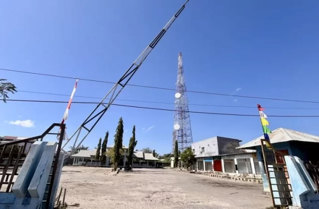 The entrance to Matawai Bus Terminal in Waingapu features a raised barrier arm, an empty dirt lot, and a tall communication tower under a clear blue sky.