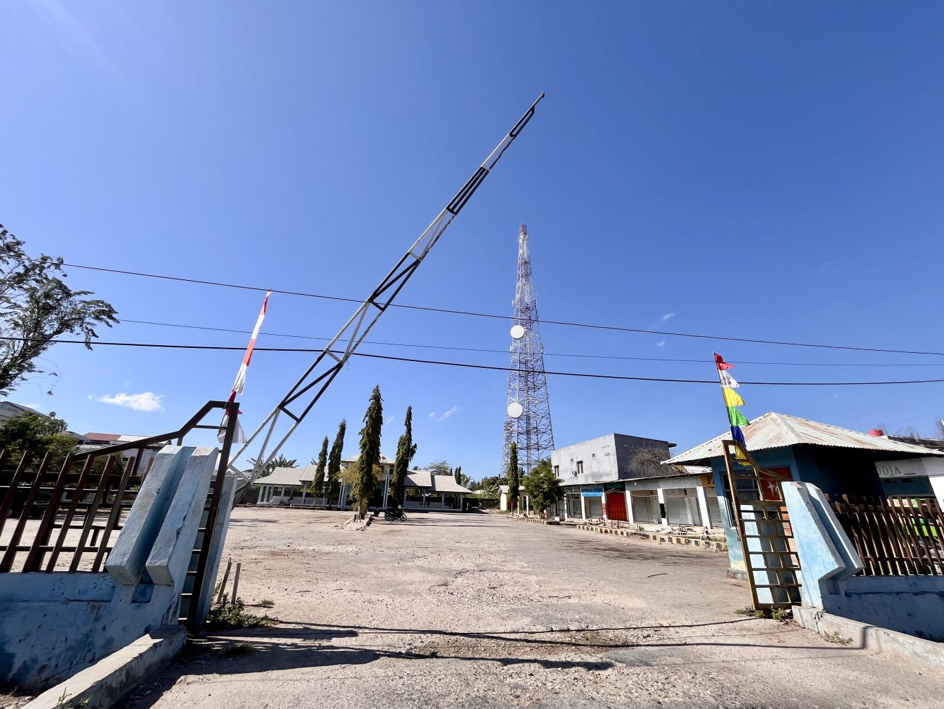 The entrance to Matawai Bus Terminal in Waingapu features a raised barrier arm, an empty dirt lot, and a tall communication tower under a clear blue sky.