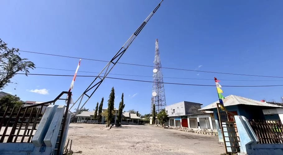 The entrance to Matawai Bus Terminal in Waingapu features a raised barrier arm, an empty dirt lot, and a tall communication tower under a clear blue sky.