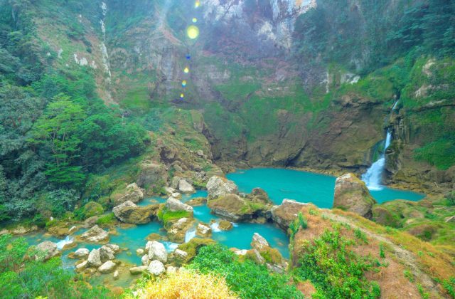 A powerful blue waterfall cascades into a large teal pool surrounded by lush green, rocky cliffs and dense rainforest vegetation in Sumba.