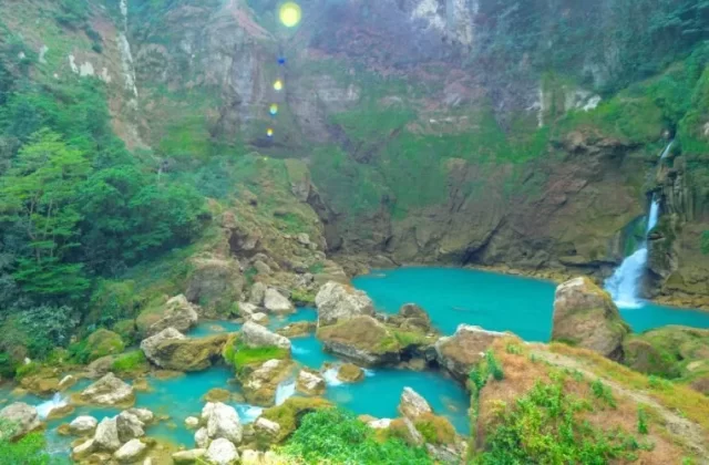 A powerful blue waterfall cascades into a large teal pool surrounded by lush green, rocky cliffs and dense rainforest vegetation in Sumba.