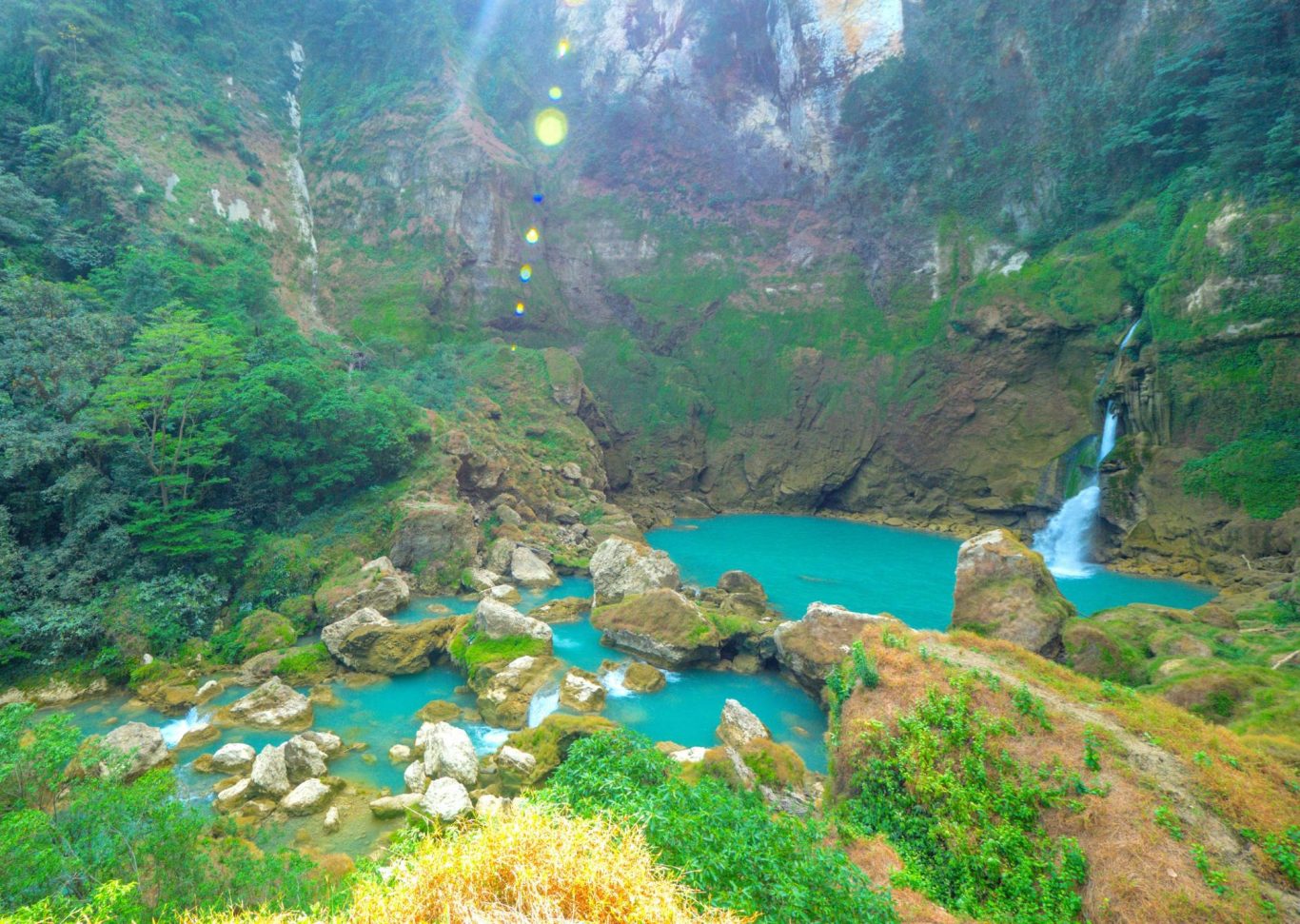 A powerful blue waterfall cascades into a large teal pool surrounded by lush green, rocky cliffs and dense rainforest vegetation in Sumba.