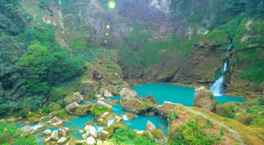 A powerful blue waterfall cascades into a large teal pool surrounded by lush green, rocky cliffs and dense rainforest vegetation in Sumba.