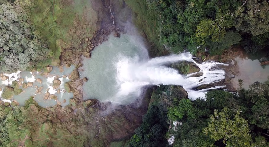 An aerial view shows the powerful Matayangu Blue waterfall plunging into a large teal pool, surrounded by lush green rainforest and rocky riverbeds.