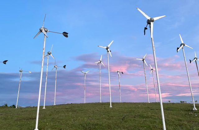 Many small, white wind turbines stand on a green grassy hilltop against a blue sky with soft pink and purple clouds.