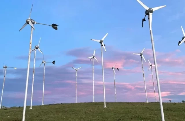 Many small, white wind turbines stand on a green grassy hilltop against a blue sky with soft pink and purple clouds.