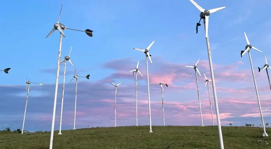Many small, white wind turbines stand on a green grassy hilltop against a blue sky with soft pink and purple clouds.