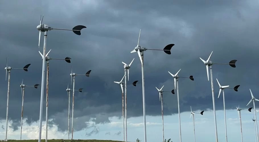 Many white wind turbines with dark blades stand on a green hilltop under a dramatic dark grey and blue cloudy sky.
