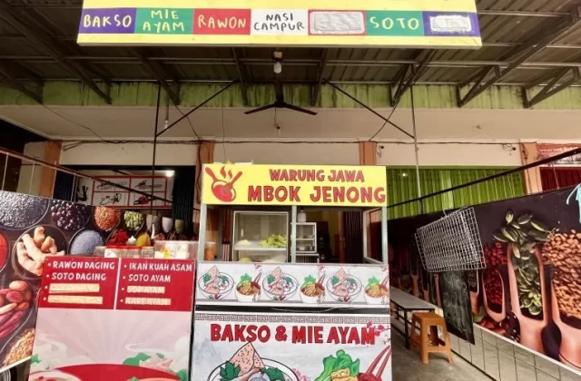 The brightly lit yellow and red 'Warung Jawa Mbok Jenong' food stall in Tambolaka displays its menu of Indonesian dishes like rawon and soto.