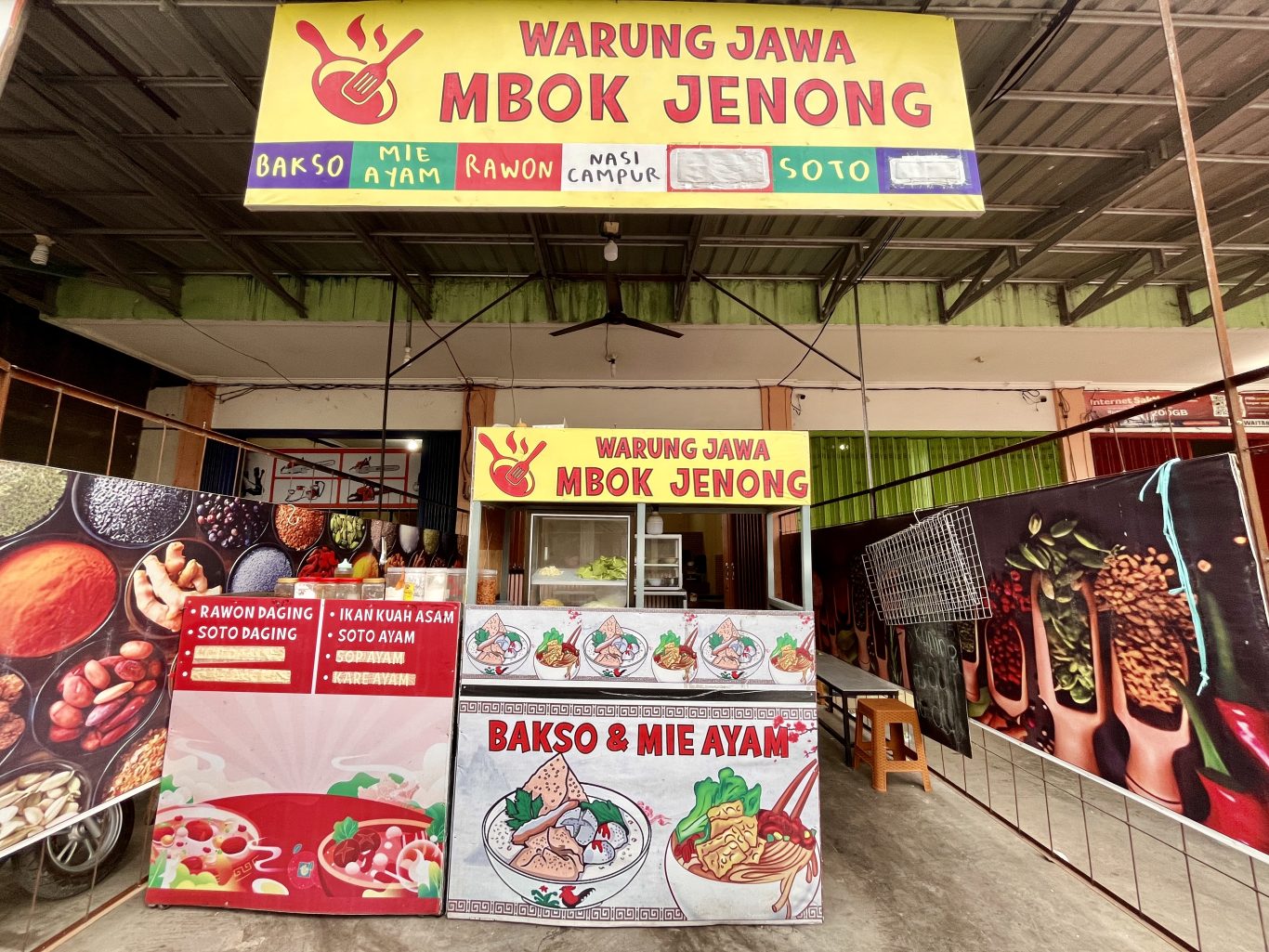 The brightly lit yellow and red 'Warung Jawa Mbok Jenong' food stall in Tambolaka displays its menu of Indonesian dishes like rawon and soto.