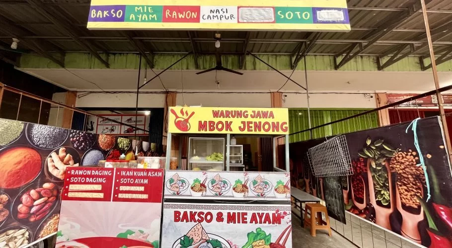 The brightly lit yellow and red 'Warung Jawa Mbok Jenong' food stall in Tambolaka displays its menu of Indonesian dishes like rawon and soto.